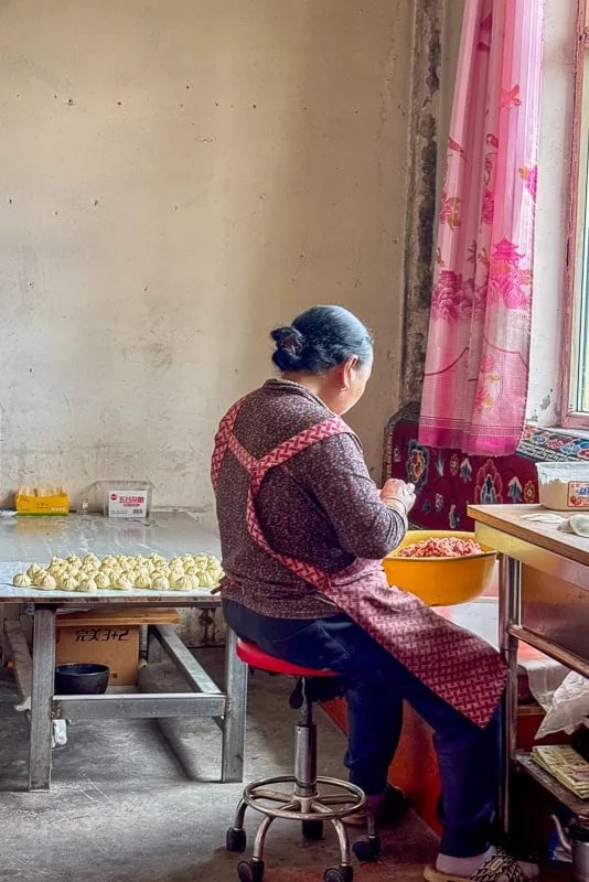 An older woman in a simple room on the Tibetan Plateau meticulously preparing many dumplings.