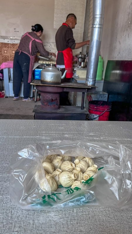 A bag of Chinese dumplings in the foreground, with two people cooking in a rustic kitchen on the Tibetan Plateau.