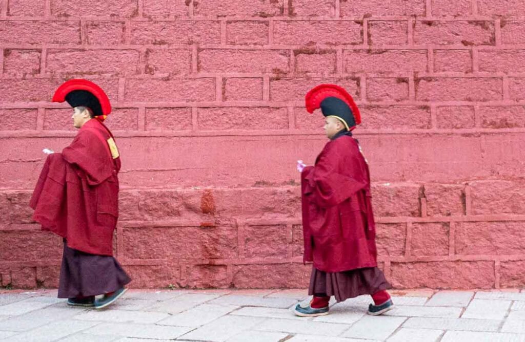 Two Tibetan monks in red robes and distinctive hats walk past a red wall in Dege, China.