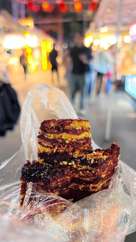 Layered red and yellow rose cake with sprinkles, held in a bag at a vibrant Xining night market.