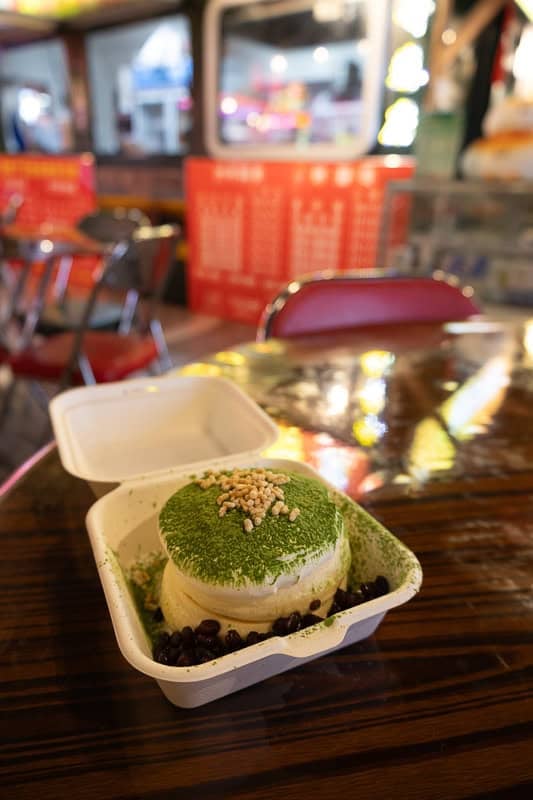 Fluffy matcha souffle pancake topped with pine nuts and red beans in a takeout container on a wooden table.