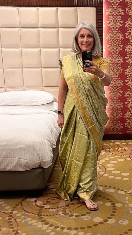 Smiling woman in a gold sari and bindi takes a mirror selfie in a hotel room.
