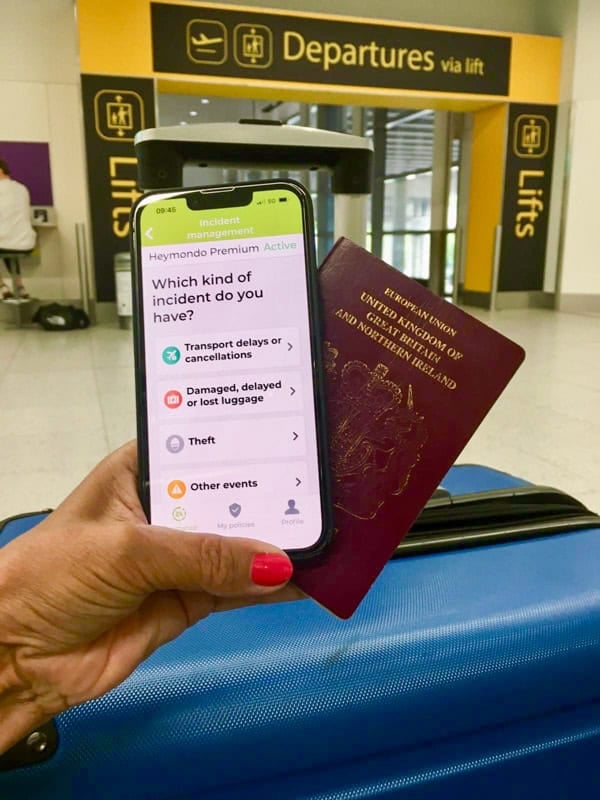 Woman's hand holding a phone with Heymondo insurance app displayed and a passport. In the background is the departures corridor and a blue suitcase.