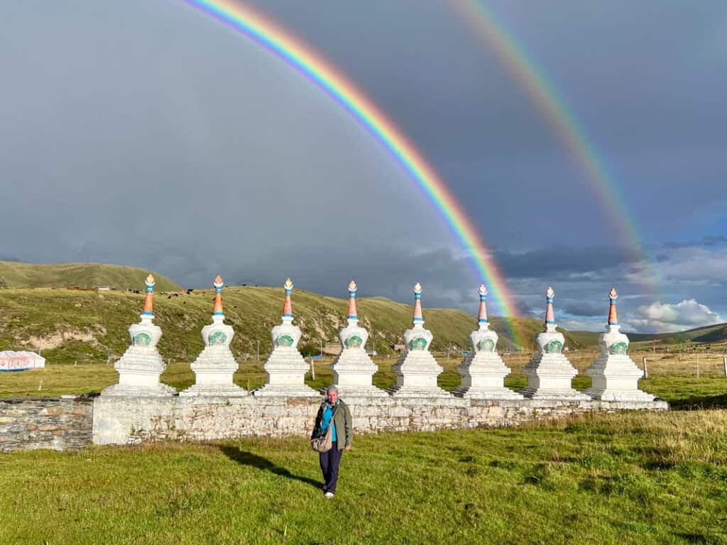 A woman standing in front of a row of stupas. In the background is a brooding grey sky and a Double rainbow