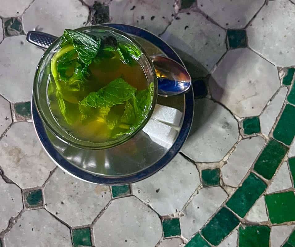 A glass of mint tea on a saucer with a spoon sits on a green and white mosaic table