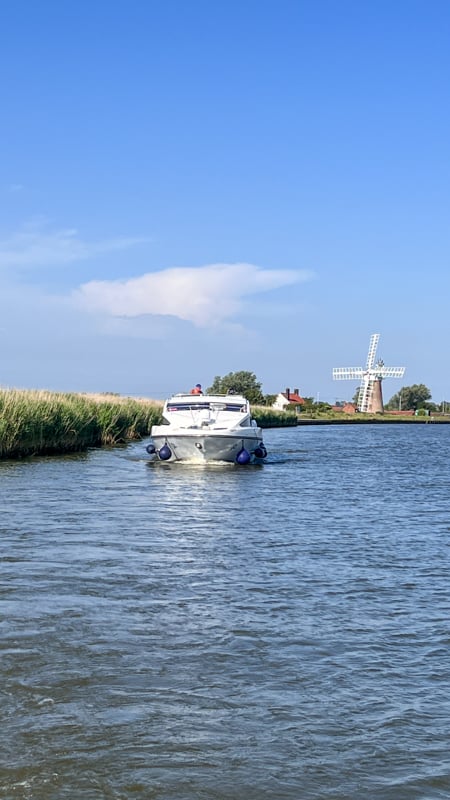 A boat on the Norfolk Broads with a windmill in the background with white sails