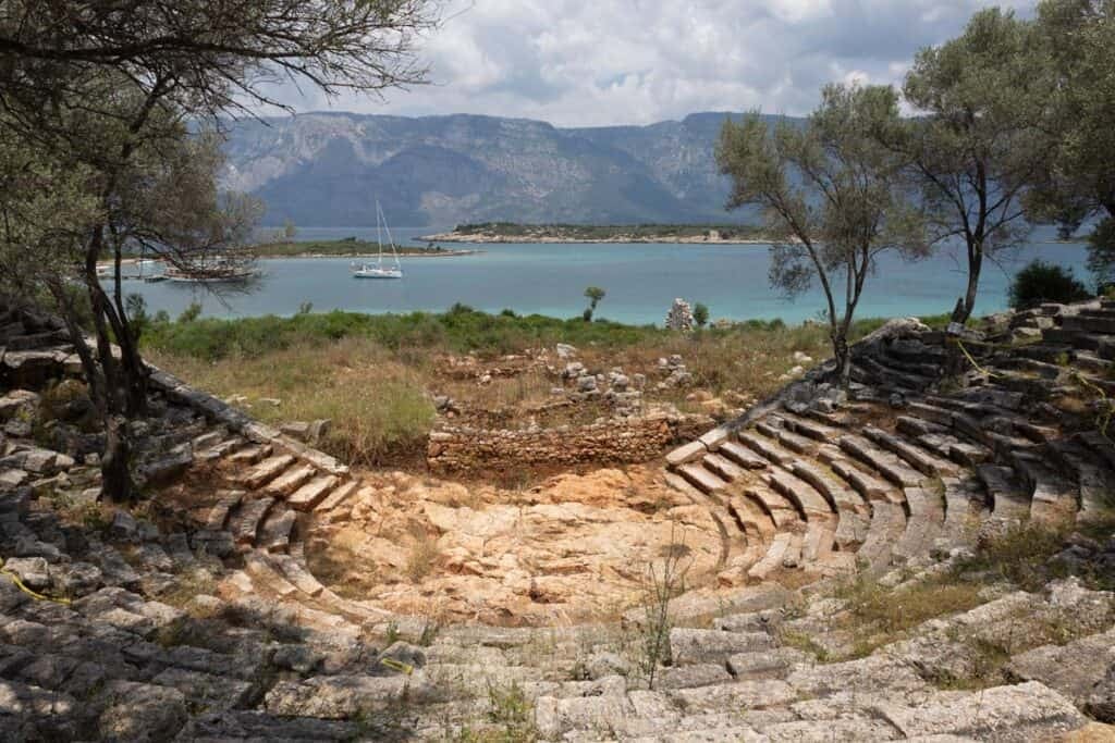 Ancient theater ruins of Aperlai, Turkey, overlook a turquoise bay with a sailboat and mountains.