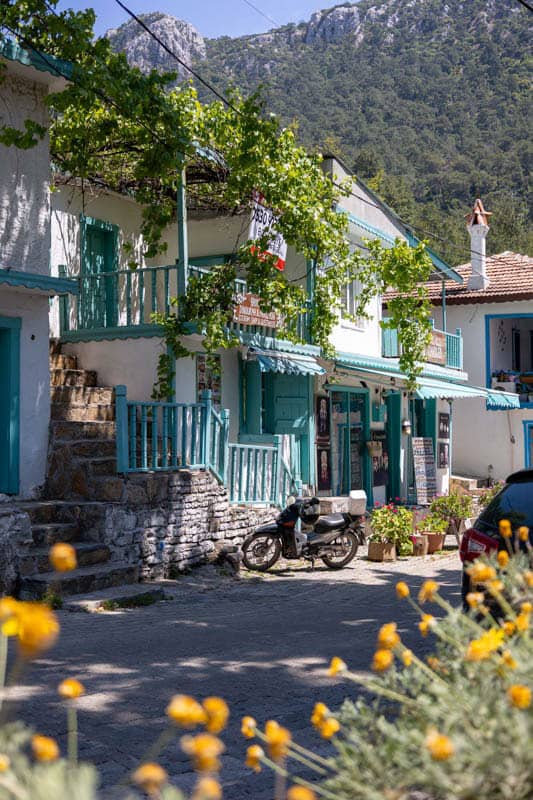 A sunny Turkish village street with white buildings, turquoise balconies, a motorcycle, and mountains in the background.