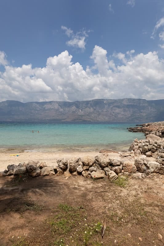 Rocky beach with clear turquoise water, two swimmers, and distant mountains under a partly cloudy sky.