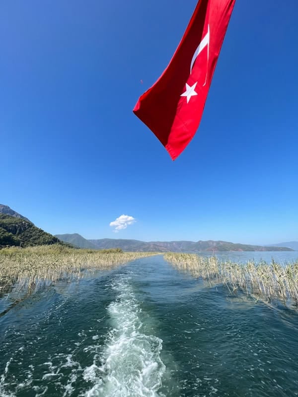 Turkish flag flying over a boat wake on the Dalyan River, Turkey, with reeds and mountains.