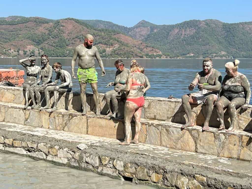 People covered in therapeutic mud at the Dalyan mud baths, with a scenic river and mountains.