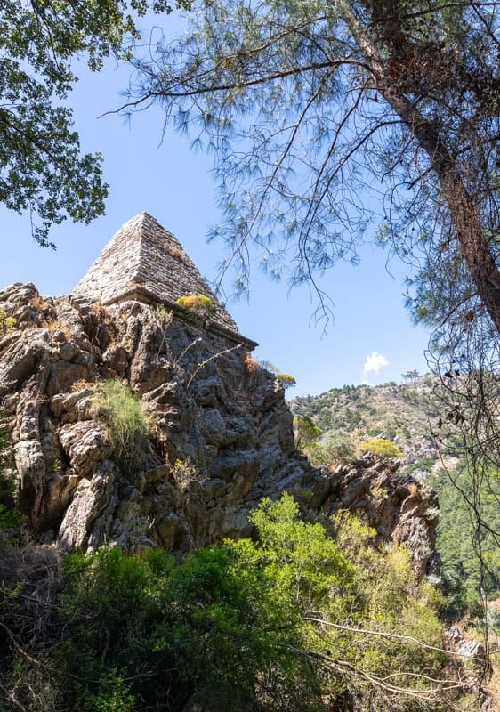 Ancient Lycian pyramidal tomb in Sidyma, Turkey, built into a rocky hillside with trees under a blue sky.