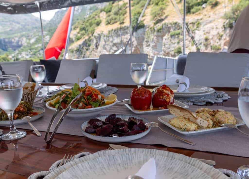 A table laid for lunch on a gulet. Stuffed peppers, garden salad, potato salad, beetroot and glasses of water are set out. A Turkish flag can be seen in the back ground.