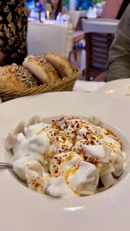 A dish of manti covered in yoghurt with honey and chilli flakes. A basket of bread is in the background