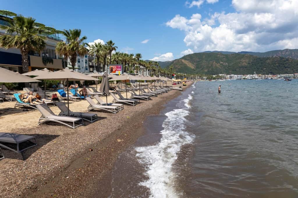 Marmaris beach with sunbeds and parasols on the gravel sand with palm trees behind. In the distance a green hills