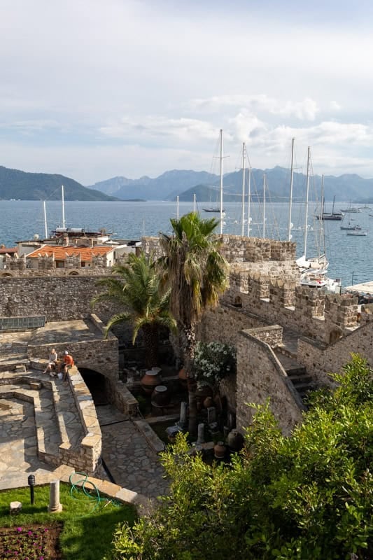Looking over the ramparts of Marmaris Castle. Palm trees and leafy trees are in the courtyard while the mast of boats can be seen behond along wth blue sea and hills in the background