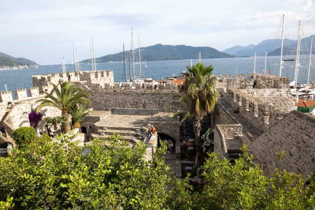 Looking over the ramparts of Marmaris Castle. Palm trees and leafy trees are in the courtyard while the mast of boats can be seen behond along wth blue sea and hills in the background