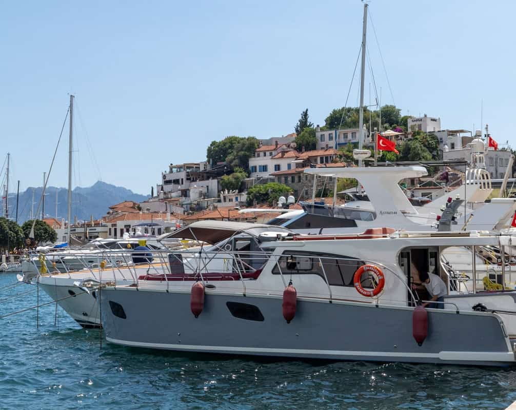 Boats are lined up on the water with Turkish flags flying form them. A small hill with buildings is in the background. The sky is blue