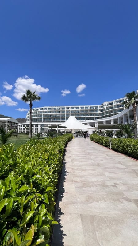 Rixos Premium Bodrum hotels curved white facade, palm trees, and paved walkway on a sunny day.