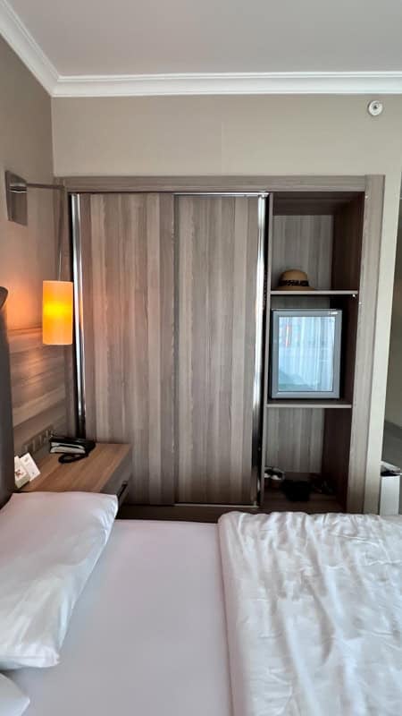 A hotel room with a white bed, a lit bedside lamp, and a wooden wardrobe with a mini-fridge and a hat.