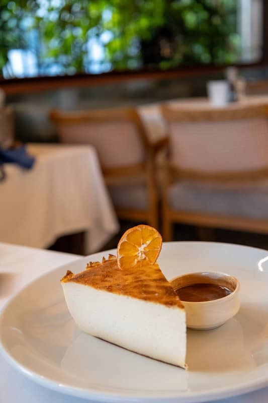 A slice of New York cheeecake garnish with a slice of dried orange sits on a plate with a small cup of chocolate sauce