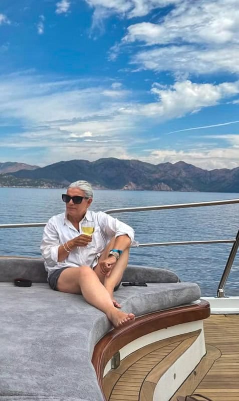 A woman wearing a white shirt and blue shorts and sunglasses sits on a gulet holding a glass of white wine. In the background is the sea and some low mountains. The sky is blue