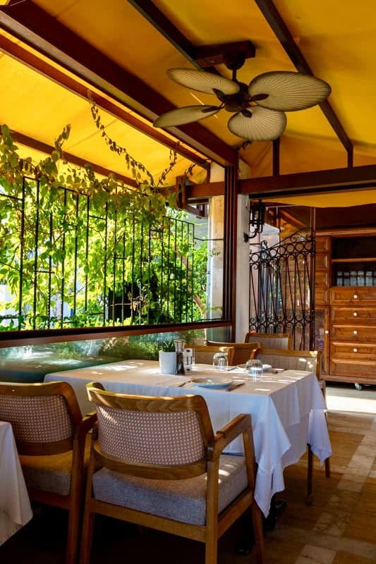 Pineapple restaurant Marmaris. A table set for four with a white cloth sits by a window. There is a fan above on the ceiling. Outside is a yellow awning and green foliage is growing outside