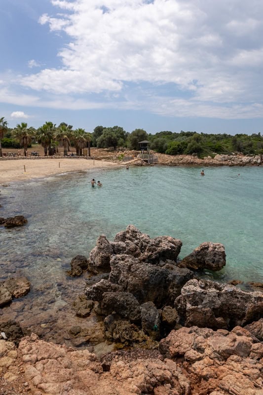 Clear turquoise water with swimmers at a sandy beach lined with palm trees and rocky outcrops under a blue sky.