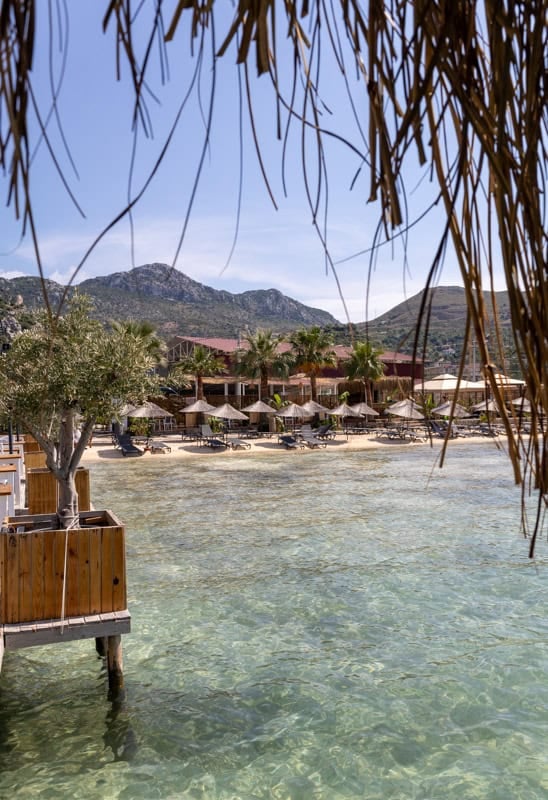 View from a thatched pier of a sunny beach with clear water, resort, and mountains in Bodrum, Turkey.