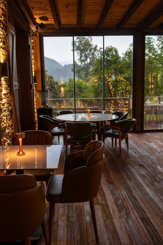 Dining tables in an ambient lit restaurant with wooden floors, floor to ceiling glass windows looking out on woodland and stone walls