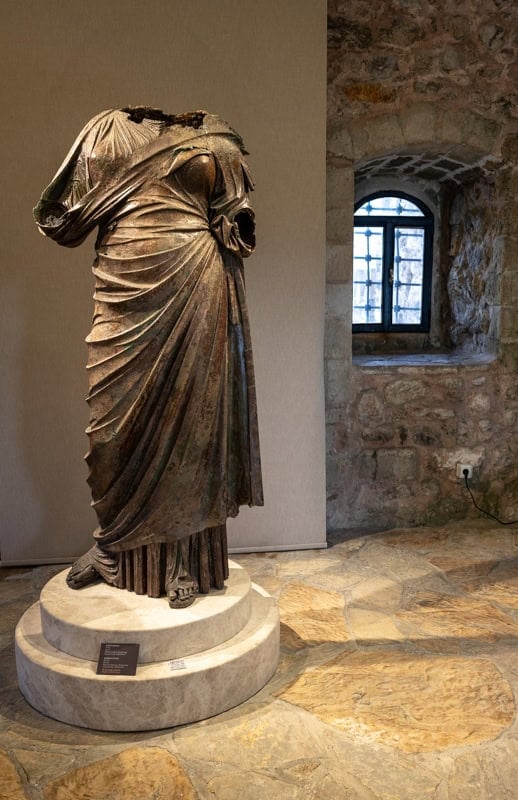 A bronze statue of a woman without arms or head stands on a low plinth in Marmaris Archaelogical Museum