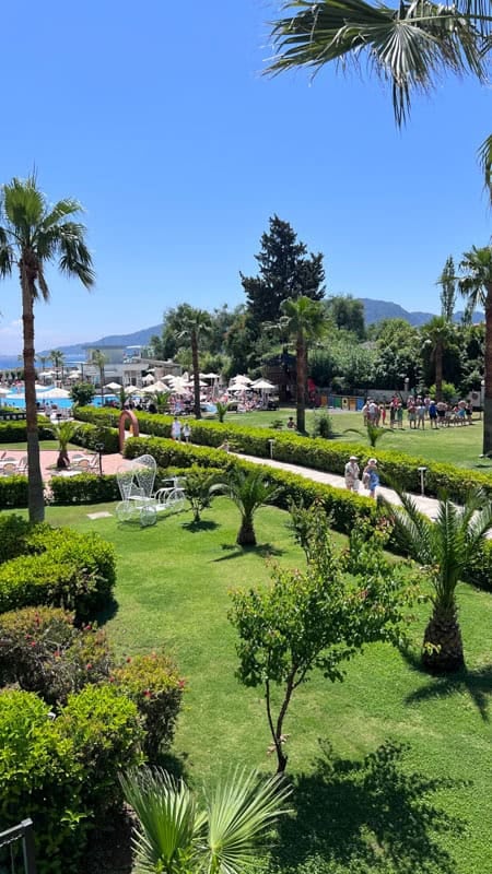 Lush resort grounds with palm trees, a swimming pool, distant mountains, and people enjoying a sunny day.