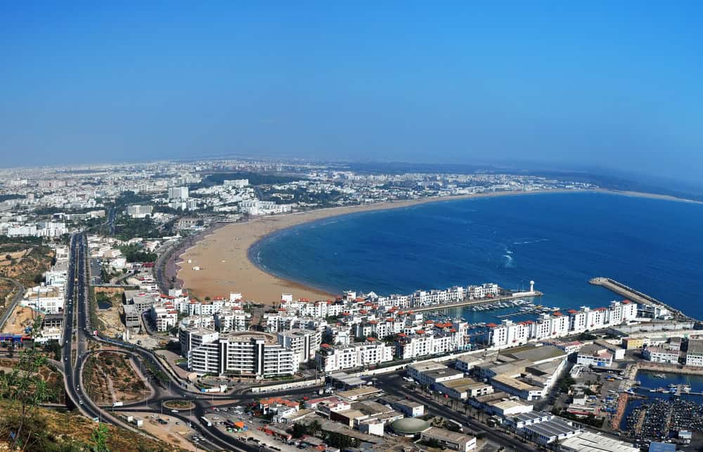 Aerial view of Agadir, Morocco, showing its crescent beach, marina, and white buildings under a clear sky.