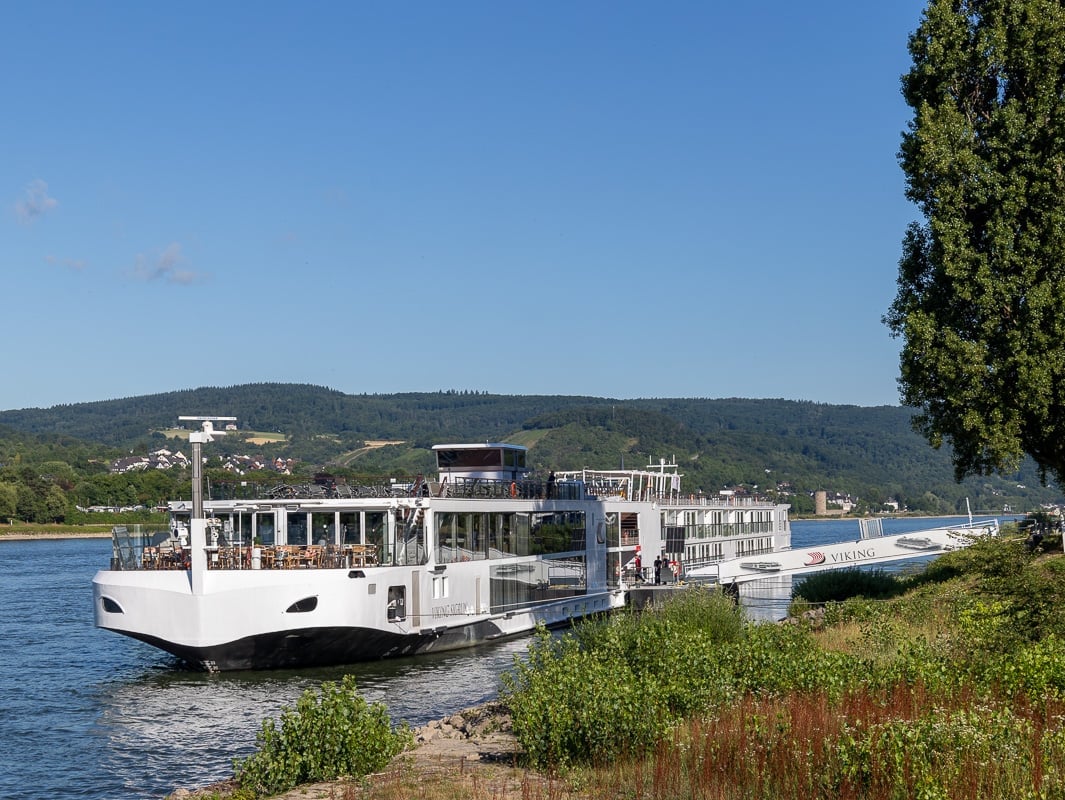 Viking Sigrun Rhine river cruise ship sits by the bank on the river Rhine. The wooded walls of the valley sit behind it and the sky is blue.